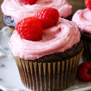 chocolate cupcakes topped with raspberry frosting, sprinkles, and raspberries on a white plate