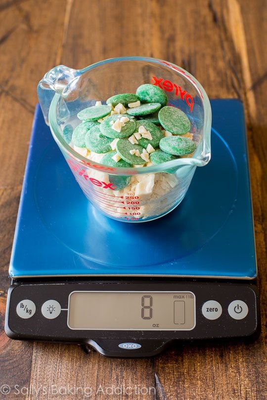 white chocolate chunks and green candy melts in a glass measuring cup on a food scale