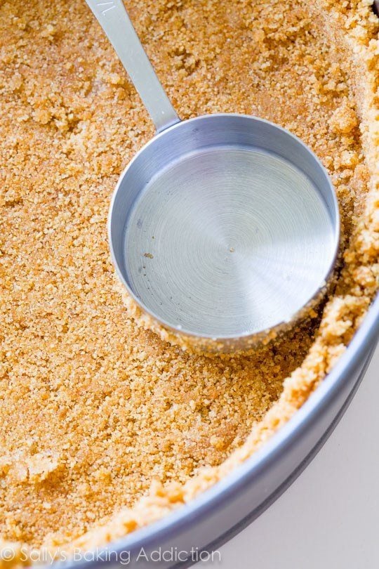 a measuring cup pressing graham cracker crust into a round baking pan
