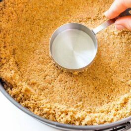 hand using a measuring cup to press graham cracker crust into a round baking pan