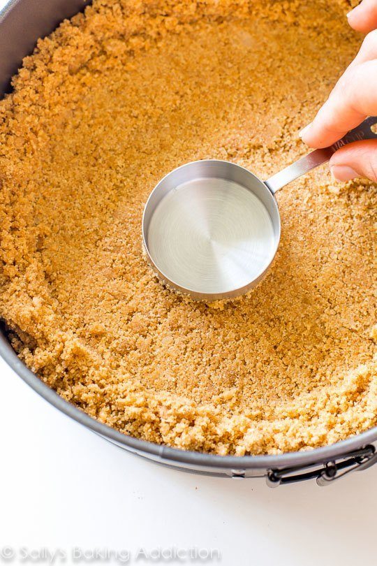 hand using a measuring cup to press graham cracker crust into a round baking pan