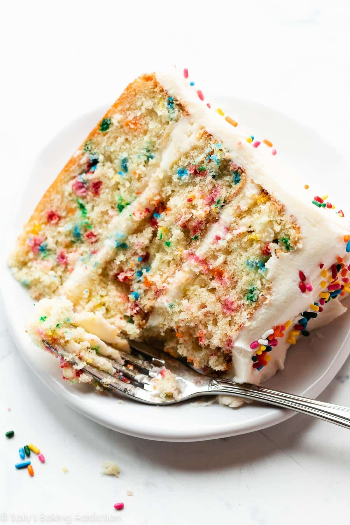 slice of Funfetti confetti birthday cake with vanilla buttercream and extra rainbow sprinkles on white plate.