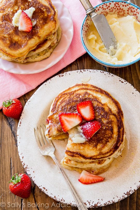 overhead image of stack of strawberry buttermilk pancakes topped with honey butter and sliced strawberries on a white plate with a fork and a bowl of honey butter