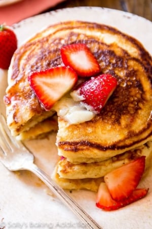 stack of strawberry buttermilk pancakes topped with honey butter and sliced strawberries on a white plate with a fork