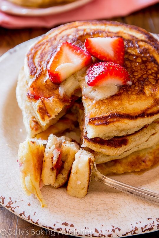 stack of strawberry buttermilk pancakes topped with honey butter and sliced strawberries on a white plate with a fork