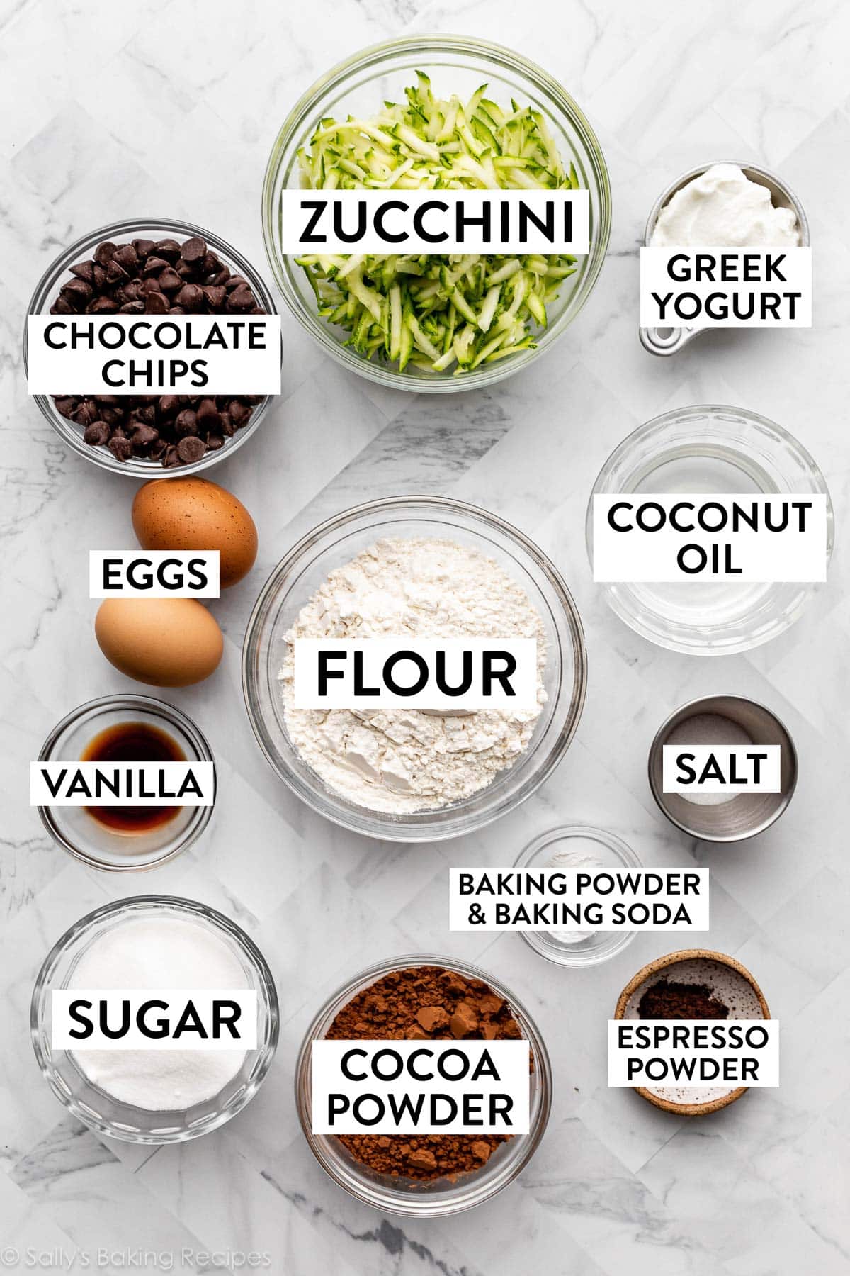 flour, coconut oil, cocoa powder, sugar, eggs, chocolate chips, and shredded zucchini in bowls on marble backdrop.