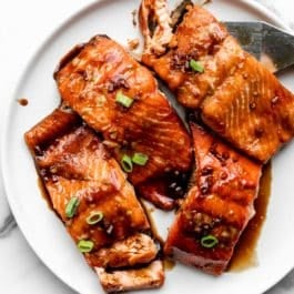 4 baked salmon filets on white plate smothered in a dark honey soy glaze and sprinkled with green onion.