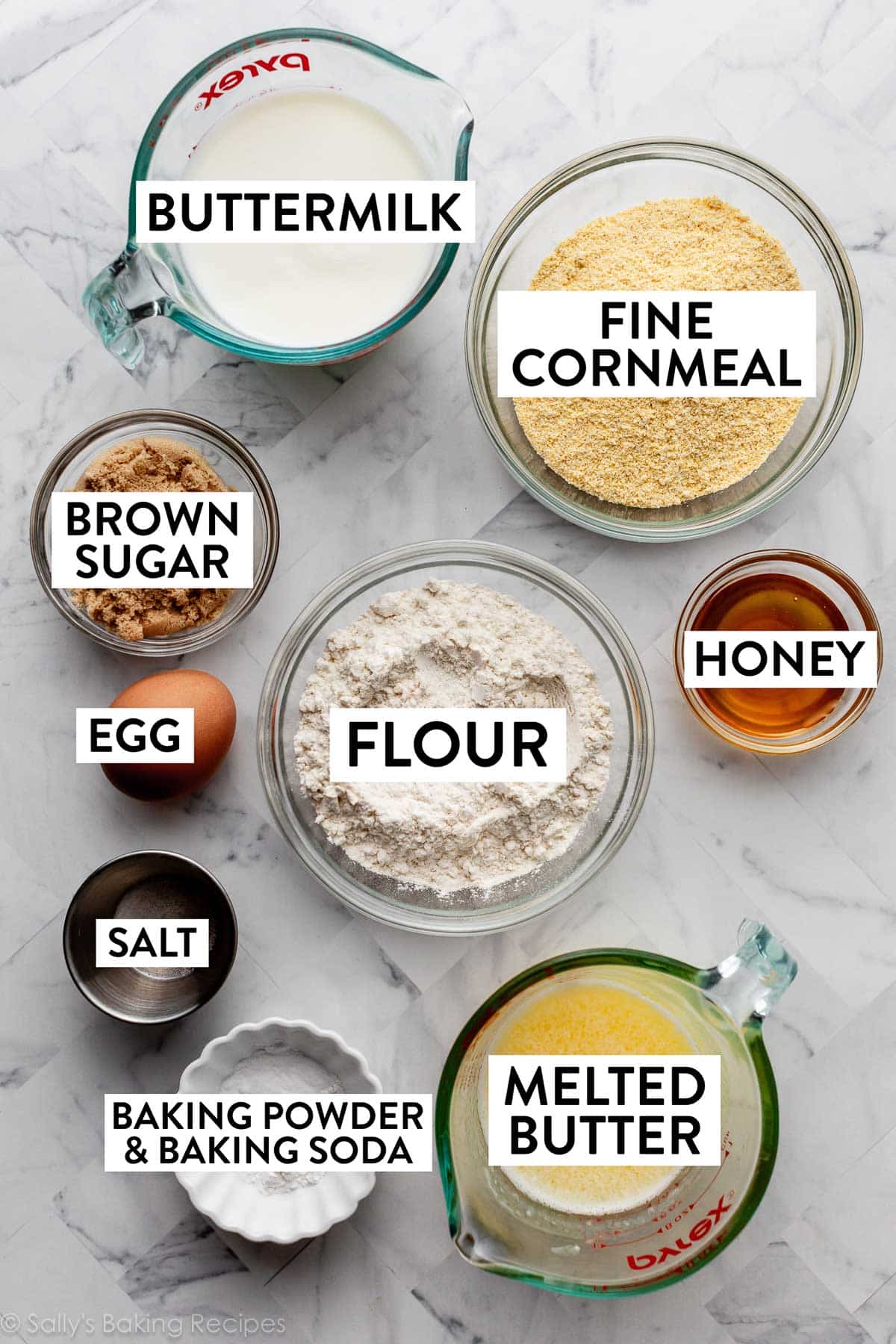 ingredients on marble counter including flour, cornmeal, buttermilk, salt, melted butter, and honey.