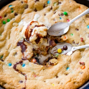 M&Ms chocolate chip skillet cookie topped with ice cream in a blue skillet with spoons