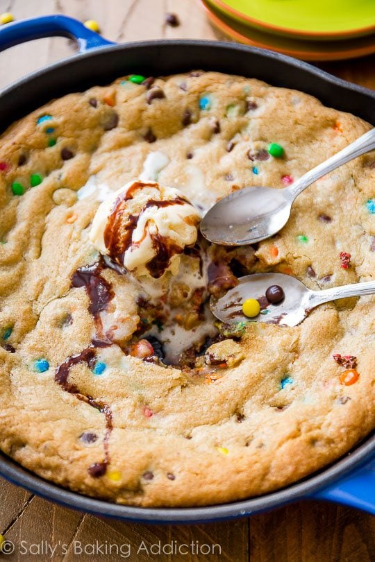 M&Ms chocolate chip skillet cookie topped with ice cream in a blue skillet with spoons