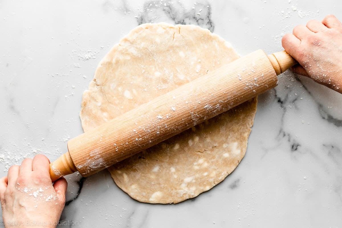 hands rolling out pie dough with wooden rolling pin on marble counter.