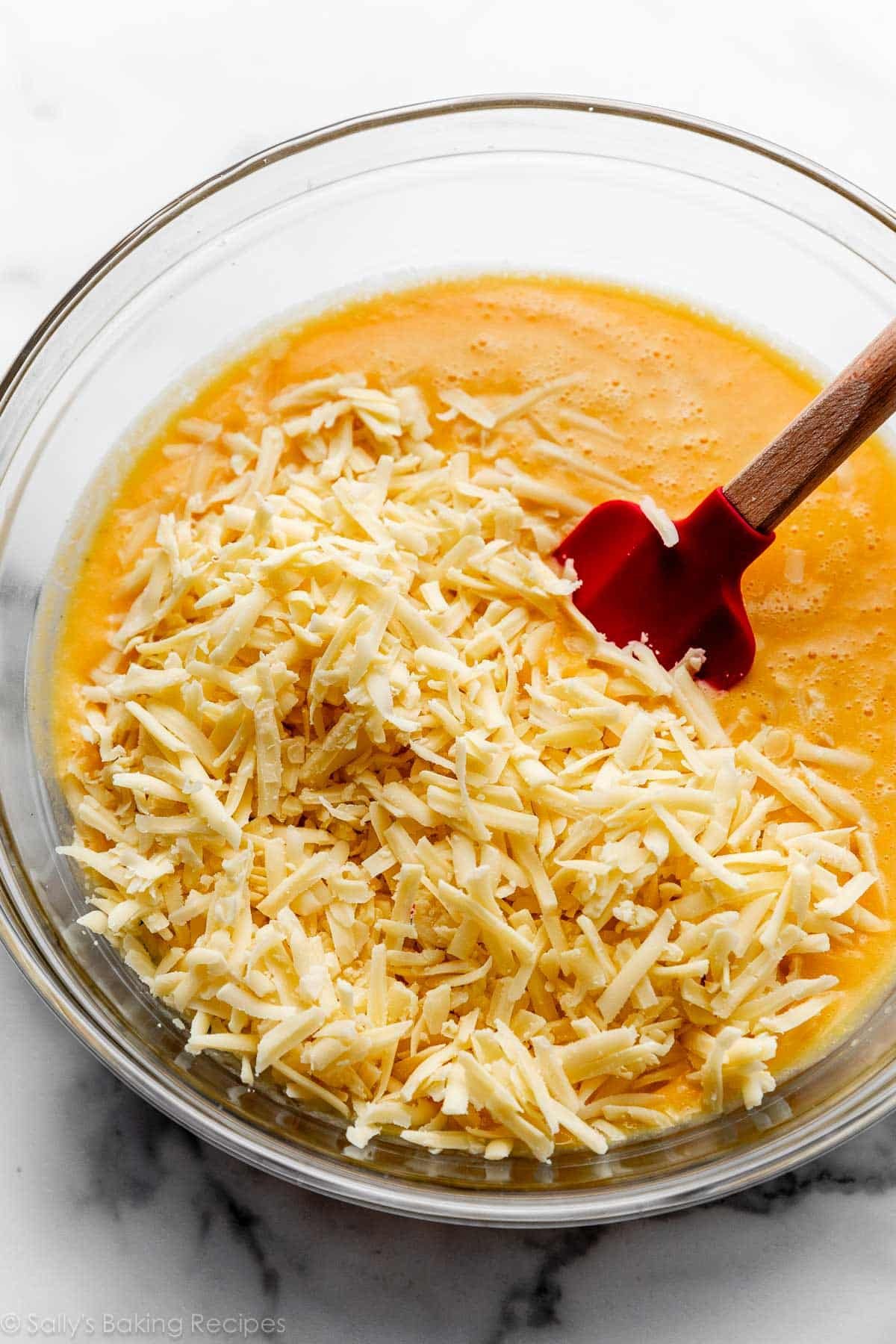 shredded gruyère cheese sitting on top of orange sauce in glass bowl with red spatula.