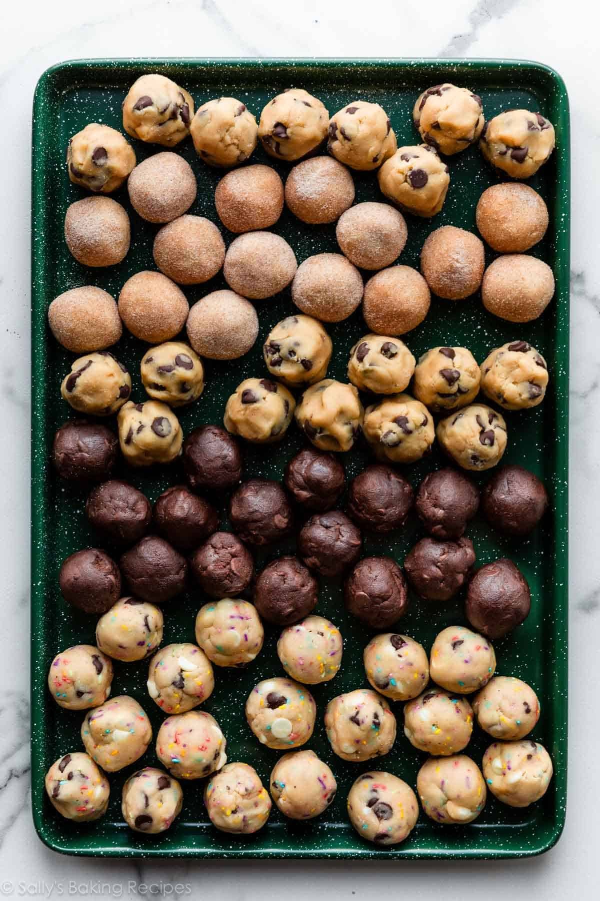 multiple varieties of frozen cookie dough balls on green baking sheet including chocolate chip cookie, chocolate chocolate chip, and snickerdoodles.