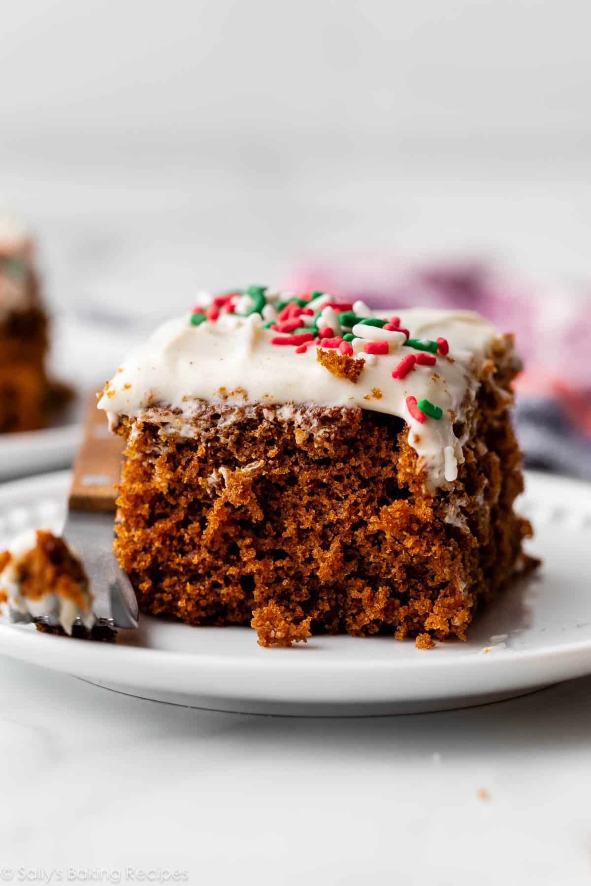 slice of gingerbread snack cake with cream cheese frosting and sprinkles on top on white plate.