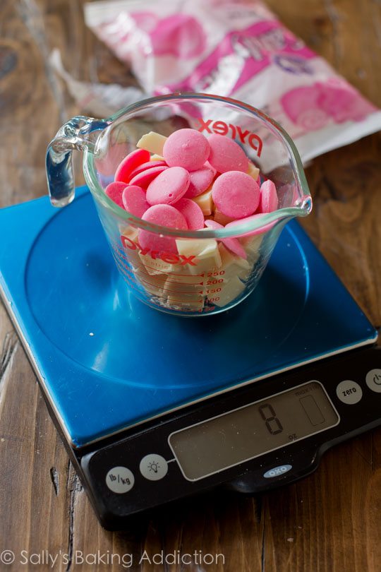 white chocolate and pink candy wafers in a glass measuring cup on a food scale