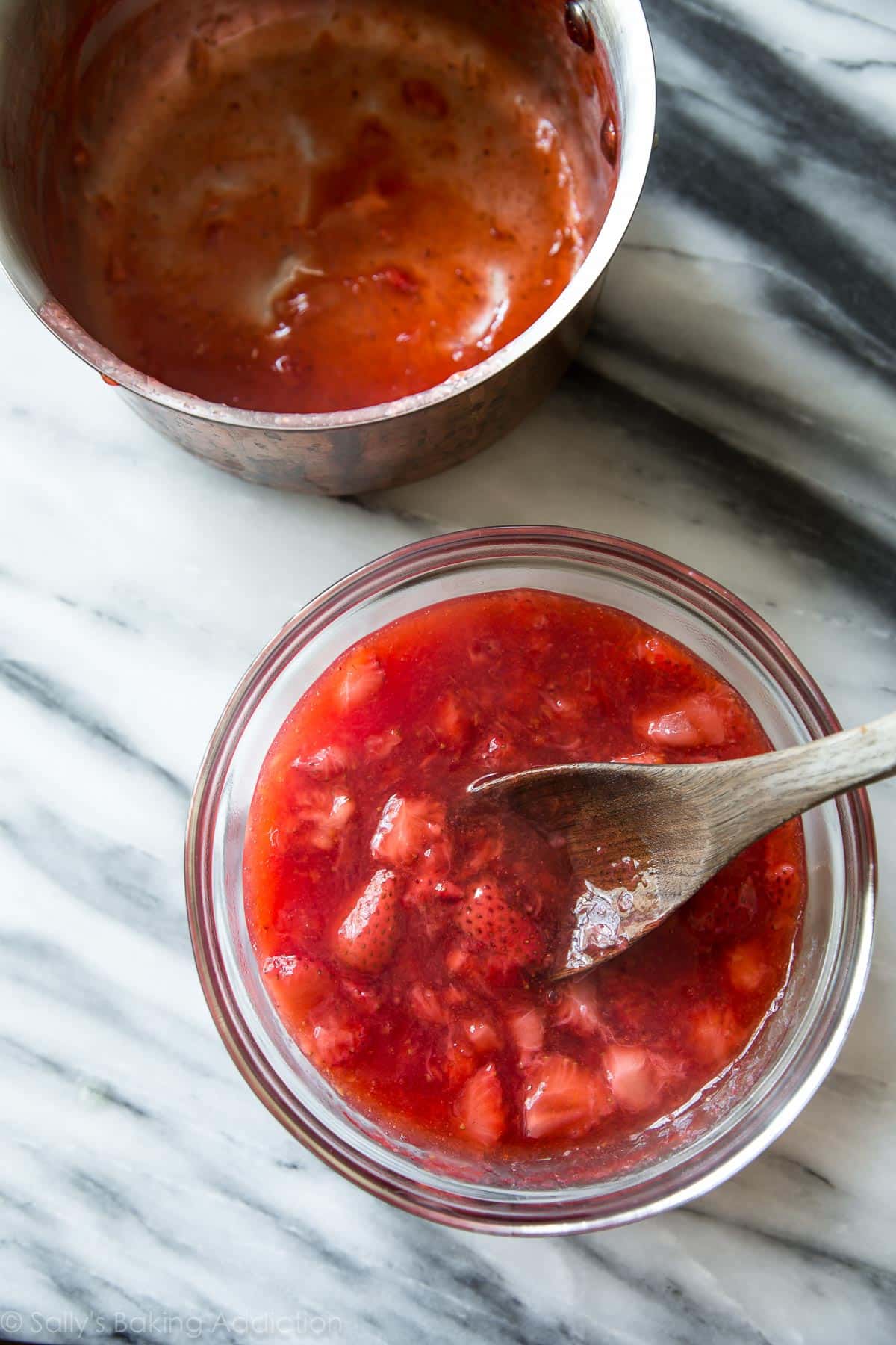 overhead image of strawberry compote in a glass bowl with a wood spoon and a saucepan
