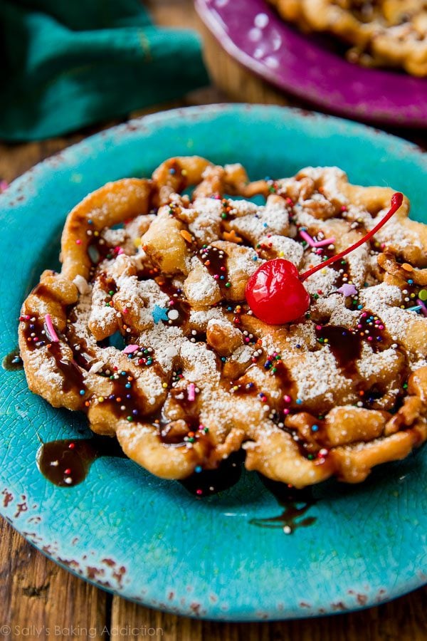 funnel cake topped with confectioners' sugar, chocolate sauce, sprinkles, and a cherry on a blue plate