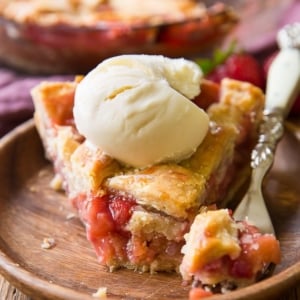 slice of strawberry rhubarb pie with a scoop of ice cream on a wood plate with a fork