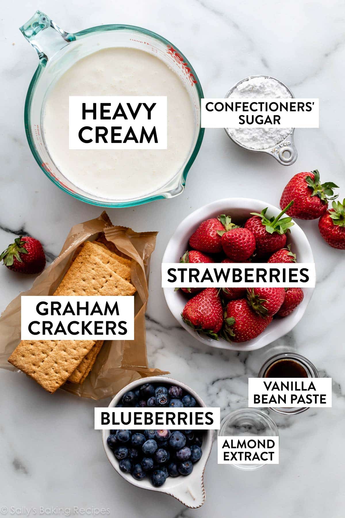 ingredients on marble counter including vanilla bean paste, graham crackers, blueberries, strawberries, heavy cream, and confectioners' sugar.