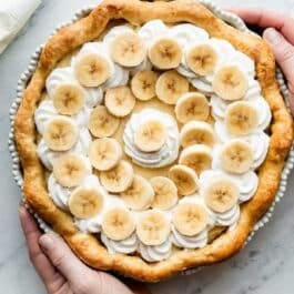hands holding a banana cream pie set on a marble countertop.