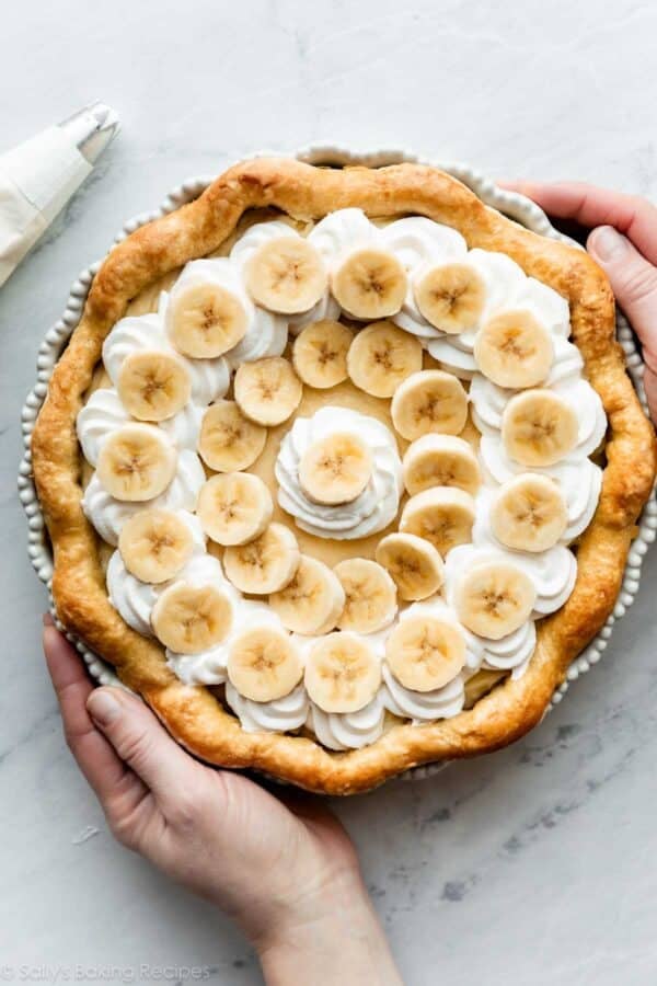 hands holding a banana cream pie set on a marble countertop.