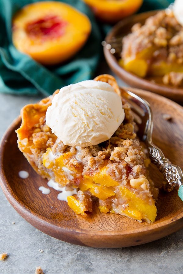 slice of peach crumble pie with a scoop of ice cream on top on a wood plate with a fork