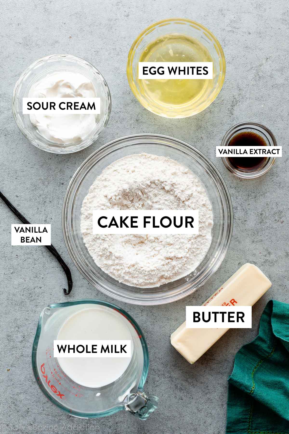 cake flour, butter, vanilla extract, milk, vanilla bean, sour cream, and egg whites on gray countertop.