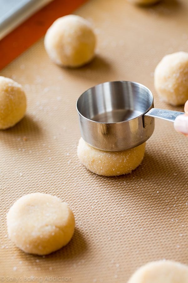 using a measuring cup to press down the tops of cream cheese sugar cookie dough balls on a silpat baking mat