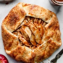apple galette with caramel sauce on white parchment paper with apples and white plates surrounding it.