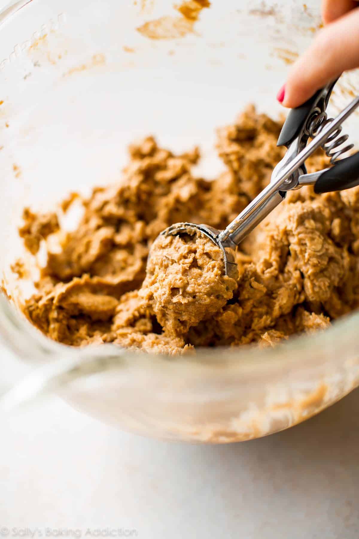 hands using a cookie scoop to scoop brown butter pumpkin oatmeal cookie dough out of a glass bowl