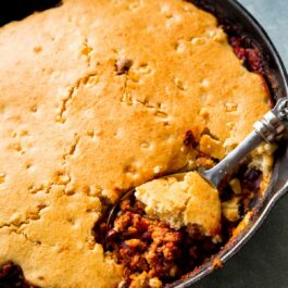 chili in a skillet topped with cornbread with a serving spoon