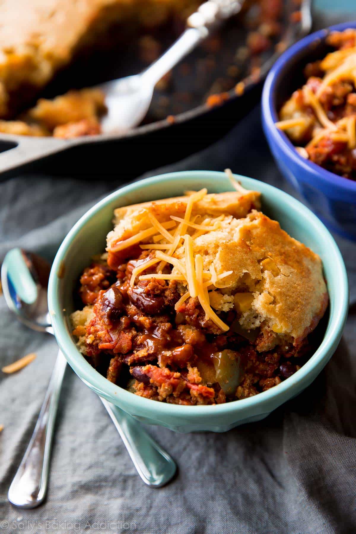 serving of cornbread topped skillet chili in a teal bowl with shredded cheese on top with a spoon