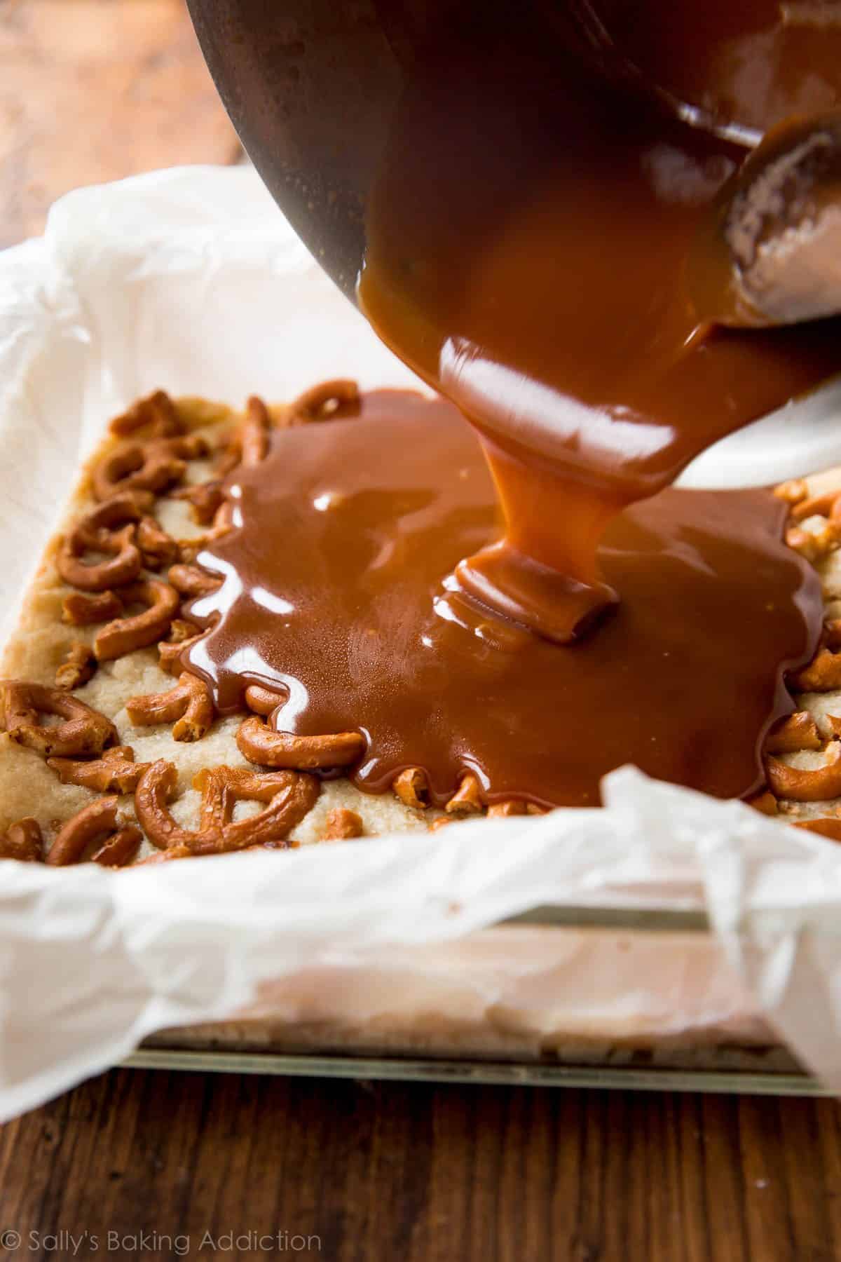 pouring salted caramel onto layer of shortbread topped with pretzels in a glass baking dish lined with parchment paper