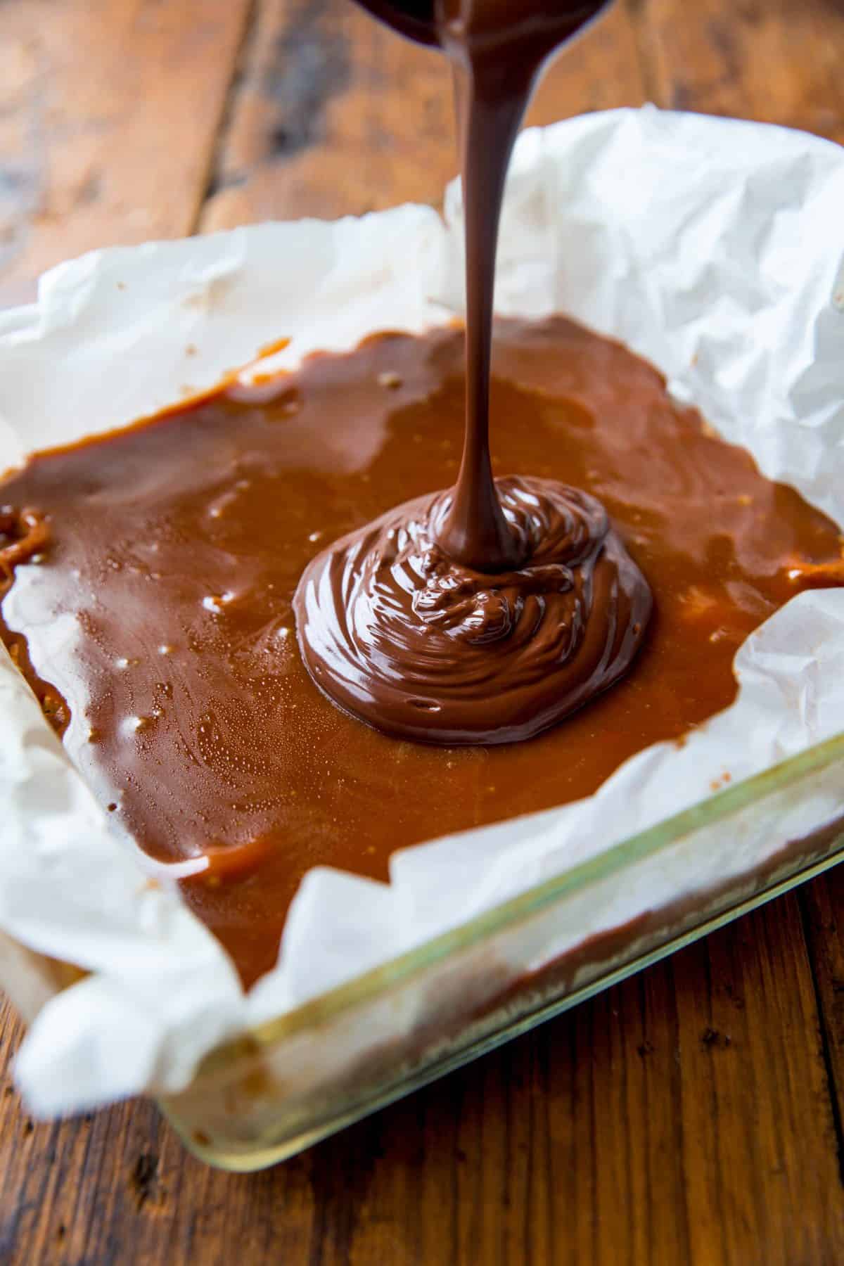 pouring melted chocolate mixture onto salted caramel layer in a glass baking dish lined with parchment paper