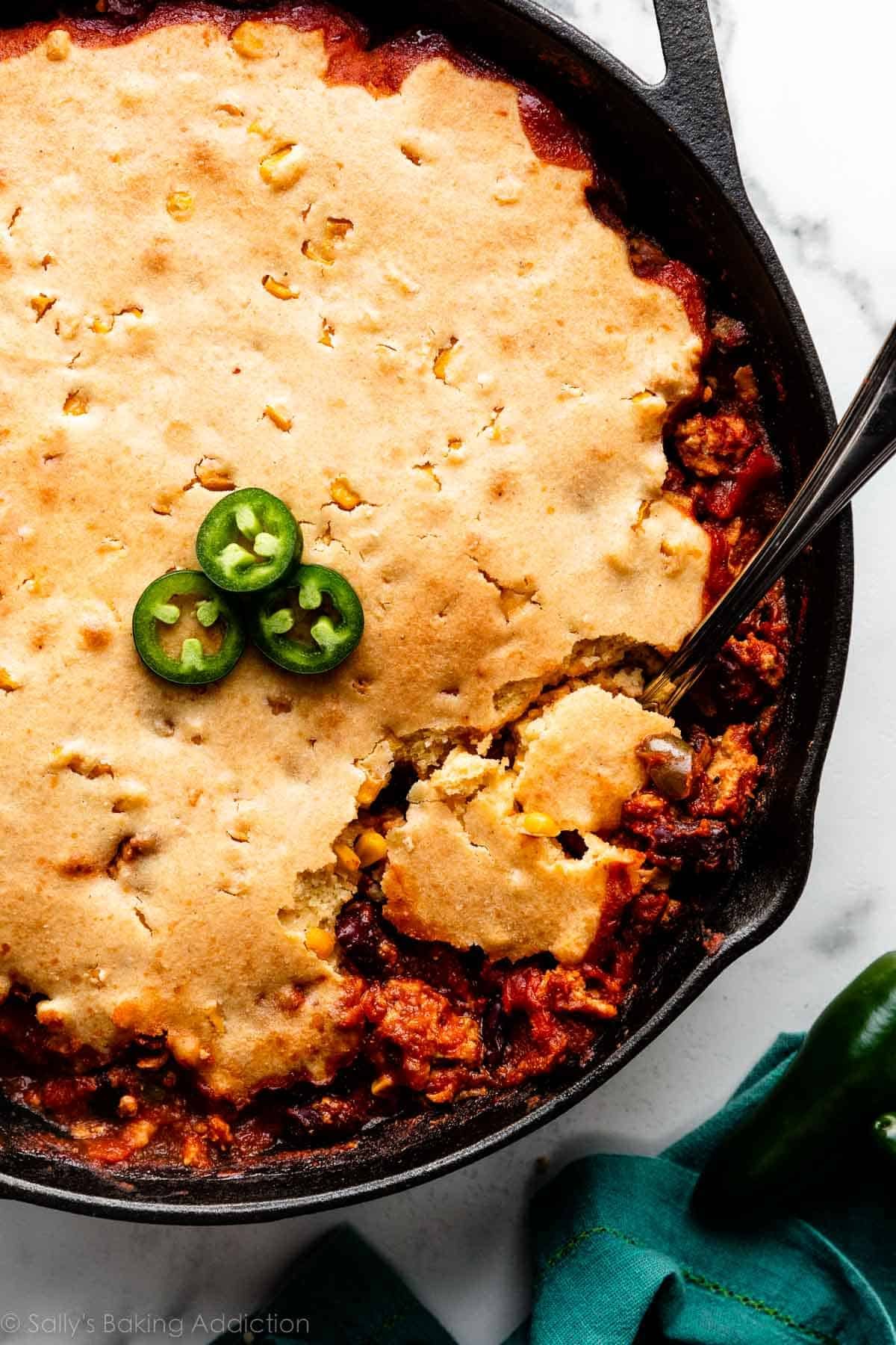 serving spoon spooning cornbread chili casserole out of a black cast iron skillet.