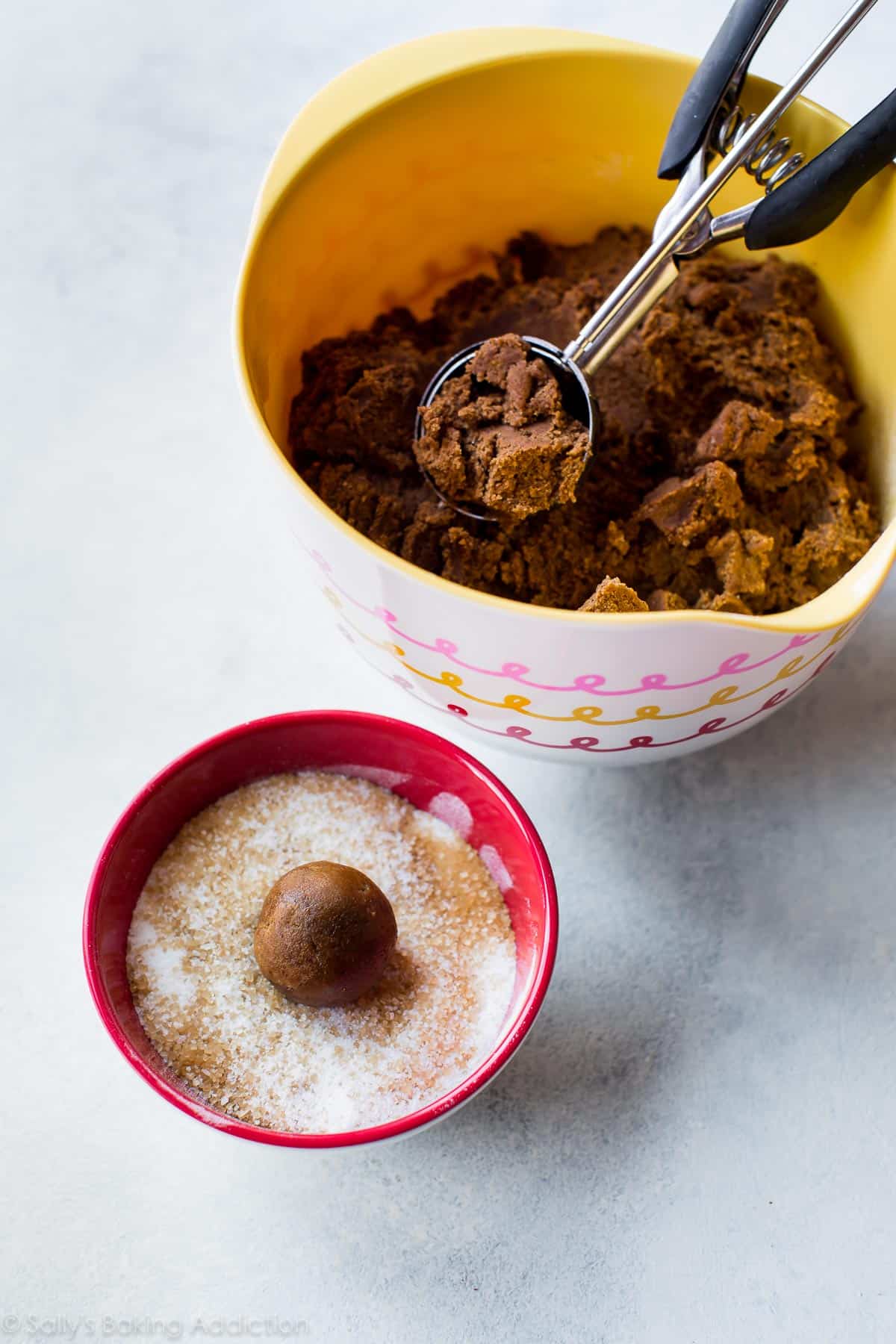 molasses cookie dough in a bowl with a cookie scoop and a cookie dough ball in a bowl of sugar for coating