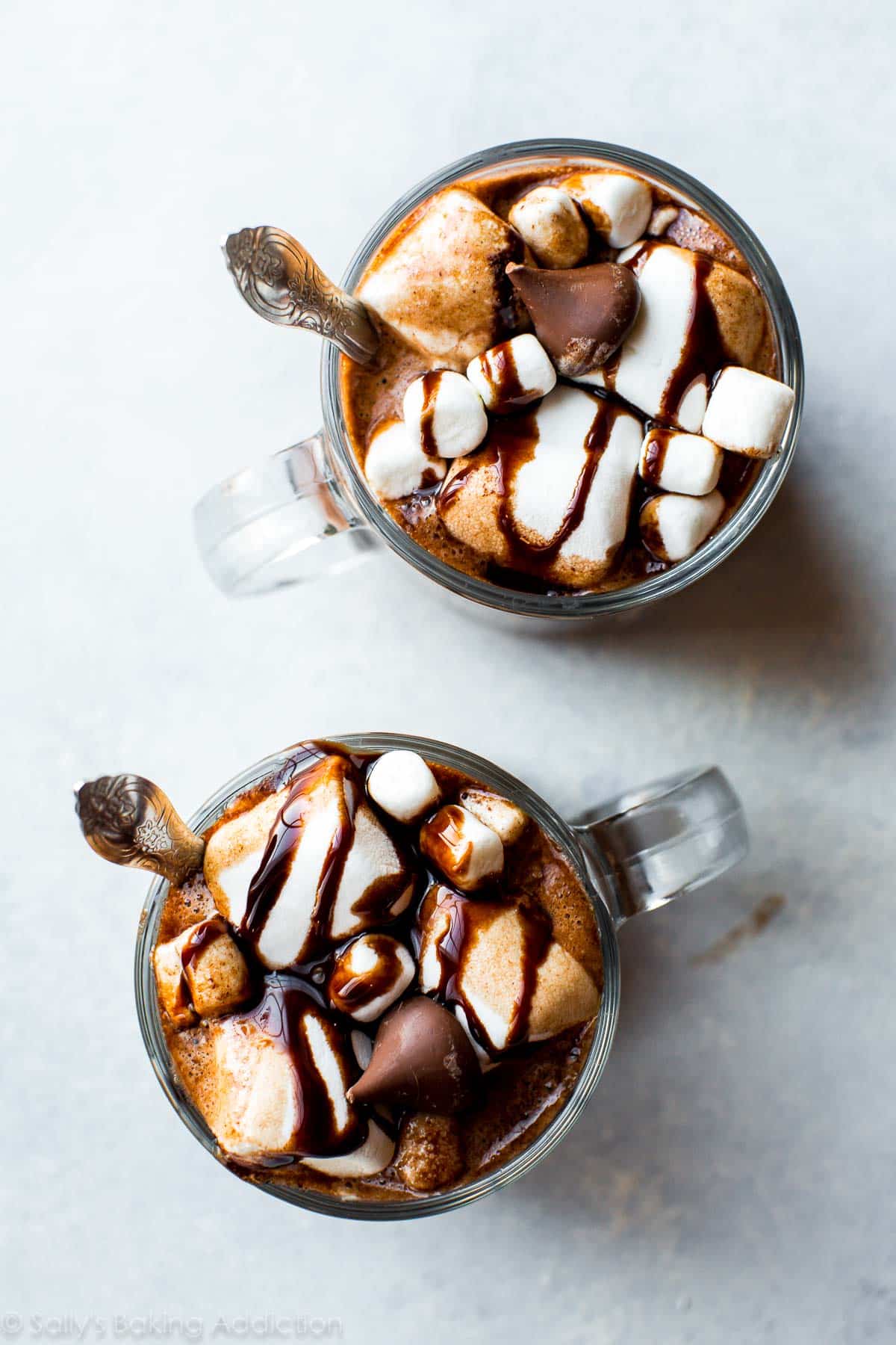 overhead image of 2 glass mugs of hot chocolate topped with marshmallows, chocolate sauce, and a Hershey's kiss