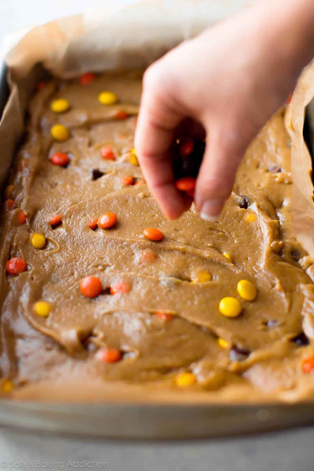 hands sprinkling Reese's Pieces onto peanut butter blondie batter in a baking pan