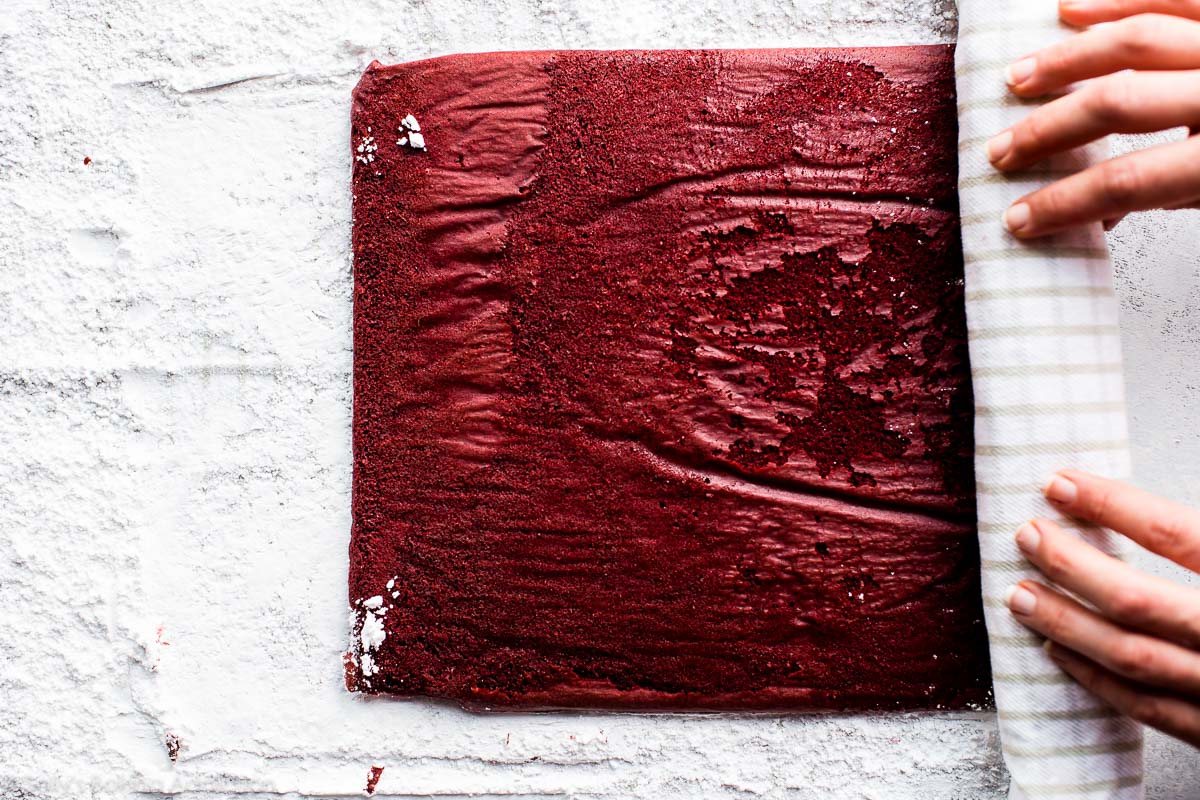 hands rolling up red velvet sponge cake into a kitchen towel