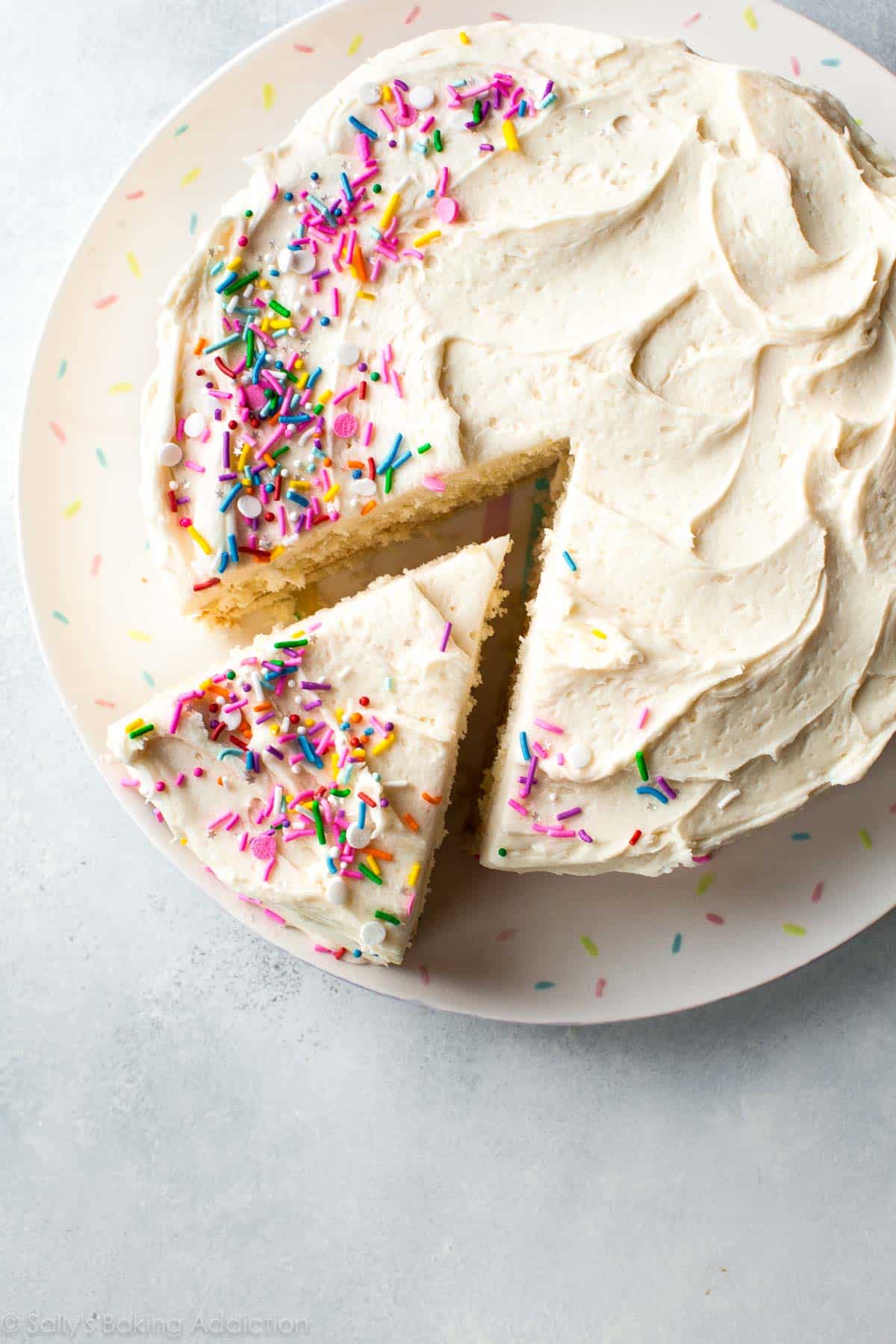 overhead image of white cake with a slice cut topped with vanilla buttercream and sprinkles