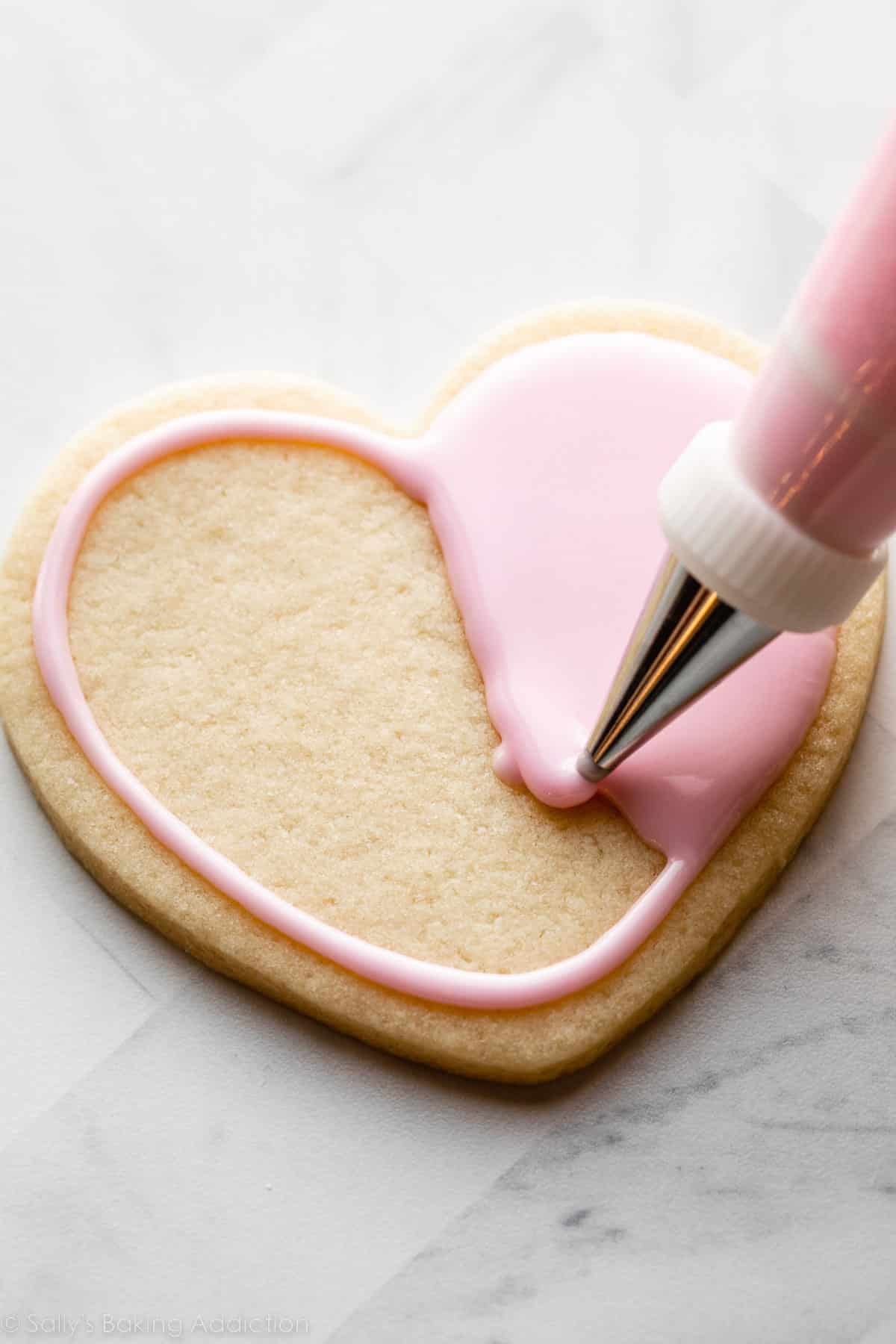 piping light pink royal icing on a heart-shaped sugar cookie.