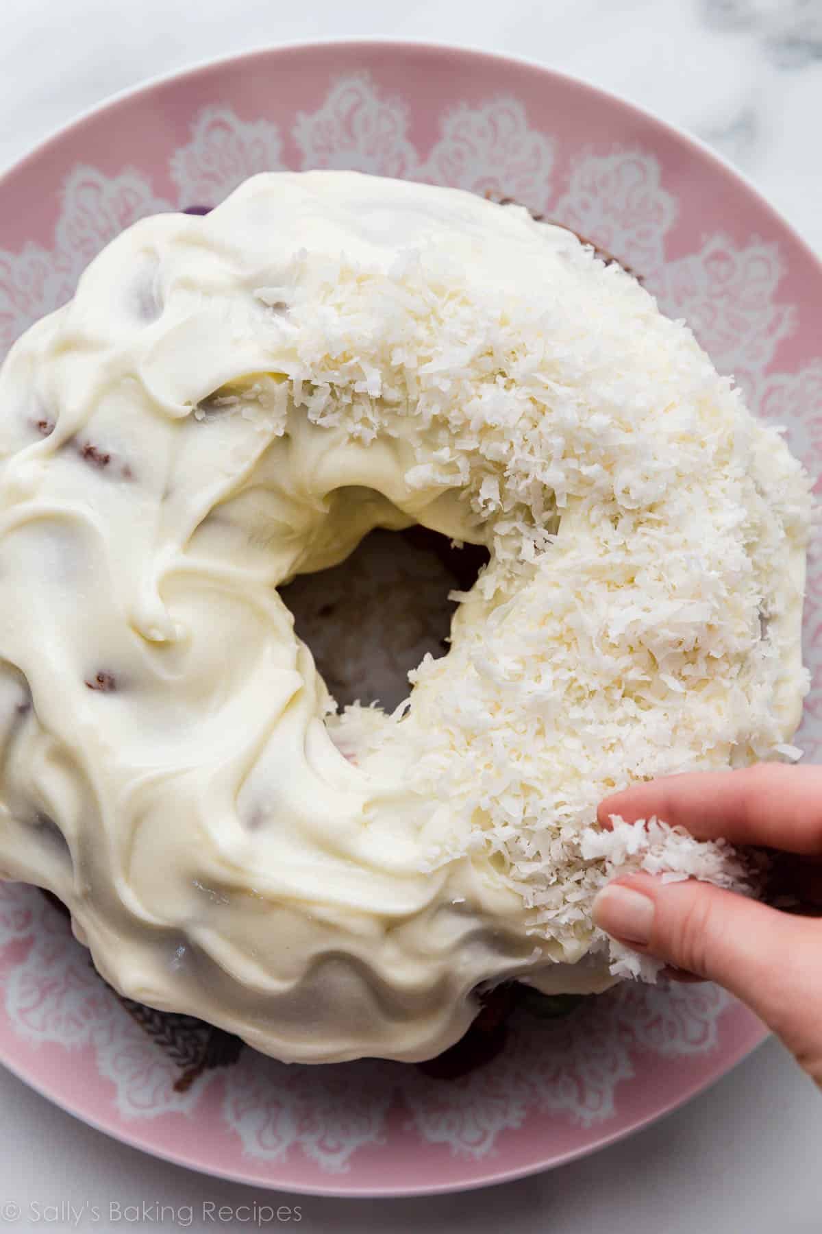 hand putting coconut on cream cheese frosted Bundt cake on top of pink serving plate.