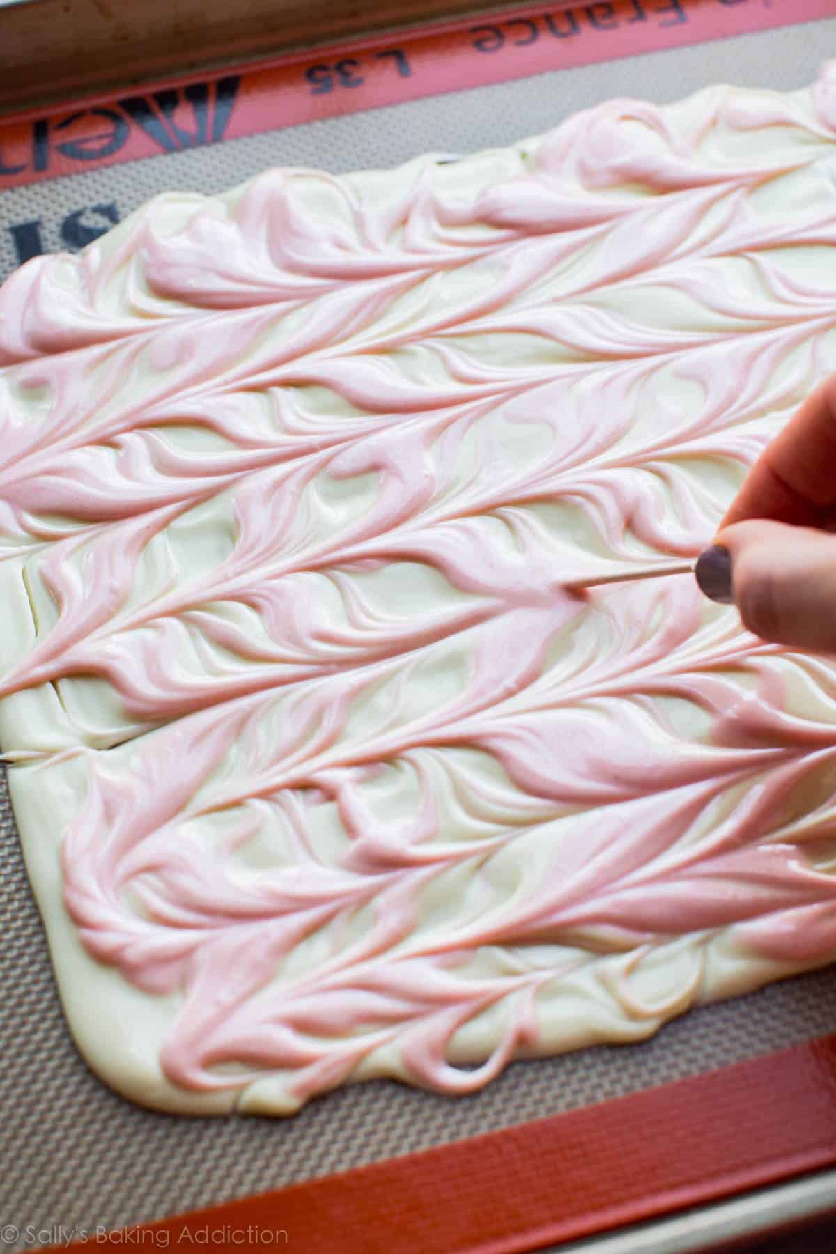 swirling white chocolate and pink colored white chocolate with a toothpick on a silpat baking mat