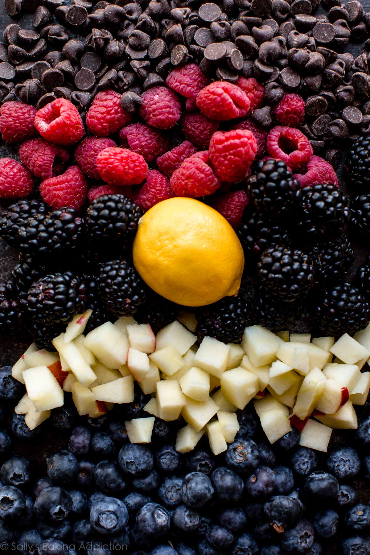 overhead image of various ingredients for muffins including chocolate chips, raspberries, blackberries, lemon, apple chunks, and blueberries