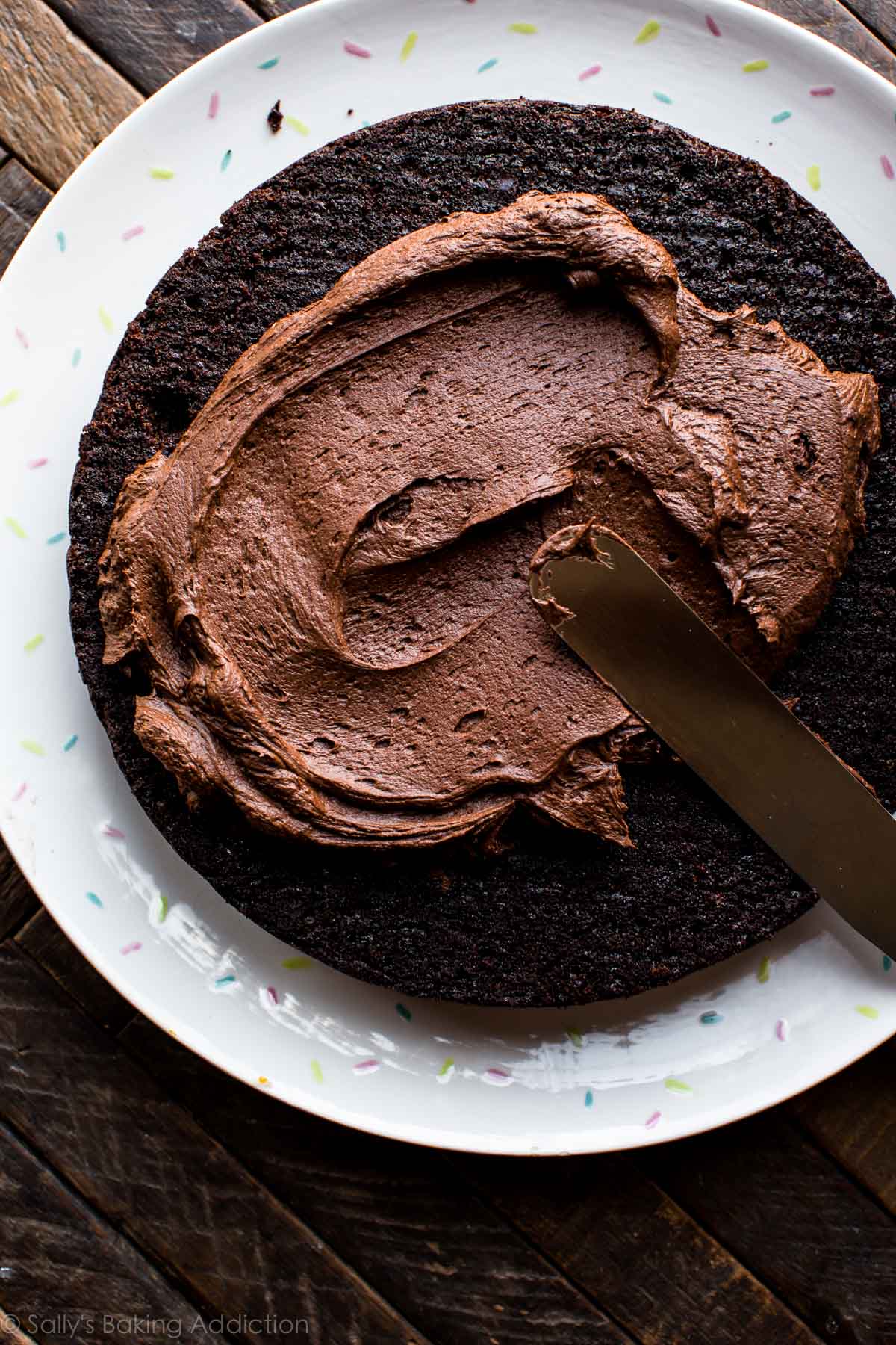 spreading chocolate fudge frosting onto layer of chocolate zucchini cake on a serving plate