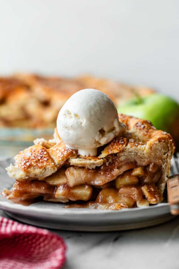 apple pie slice on plate with a scoop of vanilla ice cream on top and green apple and pie in background.