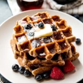 stack of whole wheat waffles on a white plate with berries, maple syrup, and butter