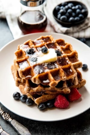 stack of whole wheat waffles on a white plate with berries, maple syrup, and butter
