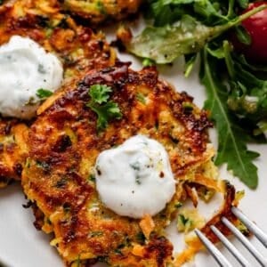 zucchini sweet potato fritter on place with yogurt sauce on top and green salad behind it.