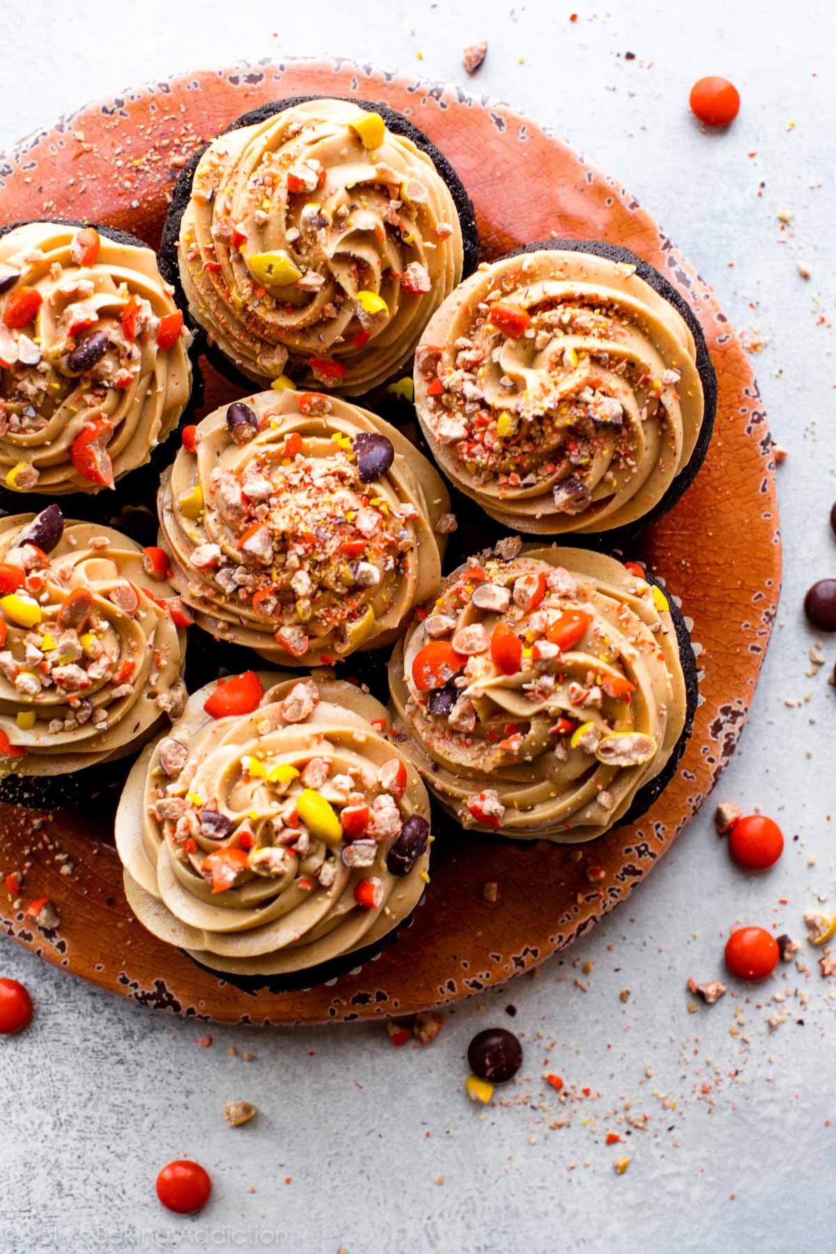 overhead image of dark chocolate cupcakes on an orange plate topped with swirls of peanut butter frosting and crushed Reese's Pieces candies